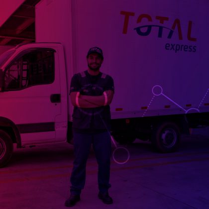 Imagem de homem sorrindo usando o uniforme da Total Express e de braços cruzados. Ao fundo um caminhão com o logo da empresa na lateral.