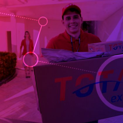 Imagem de homem com o uniforme da Total Express segurando uma caixa de papelão grande com o logotipo da empresa. Ao fundo uma mulher parada