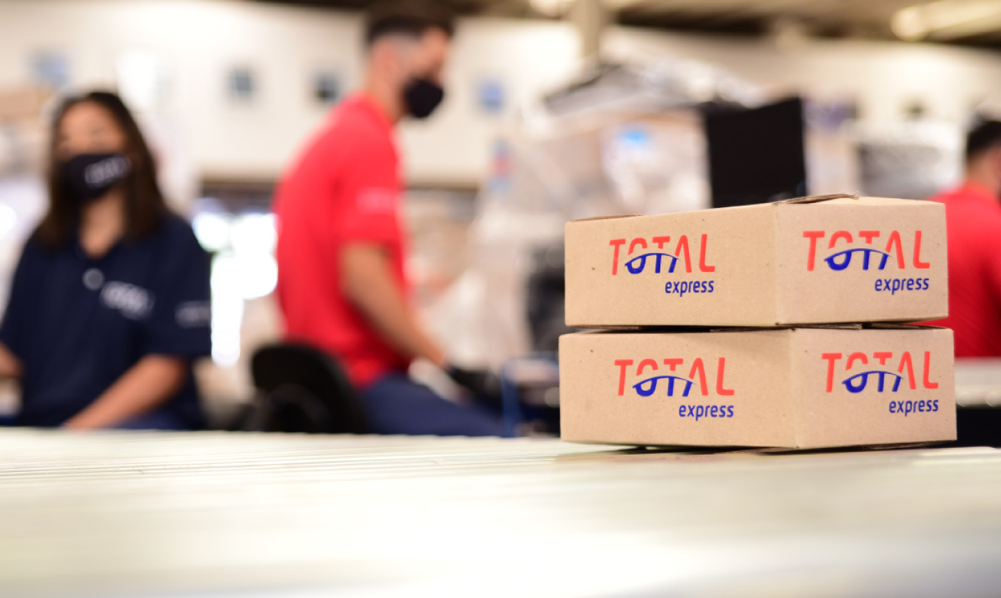 Duas caixas de papelão empilhadas, ambas com o logotipo da Total Express. Ao fundo pessoas trabalhando.