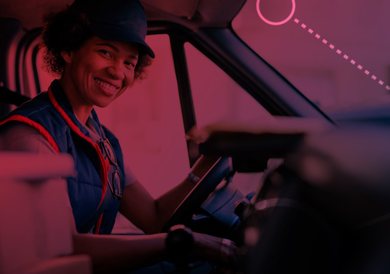 Foto de mulher sorrindo e olhando para a câmera enquanto dirige um veículo. Na frente dela aparece um volante.